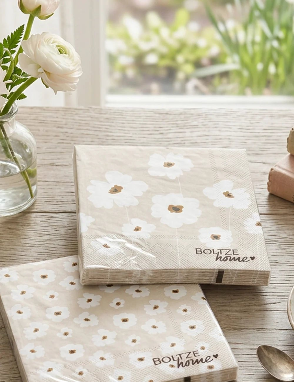 Zwei Servietten-Packungen Canea mit großem und kleinem Blumenmuster neben weißen Ranunkeln
